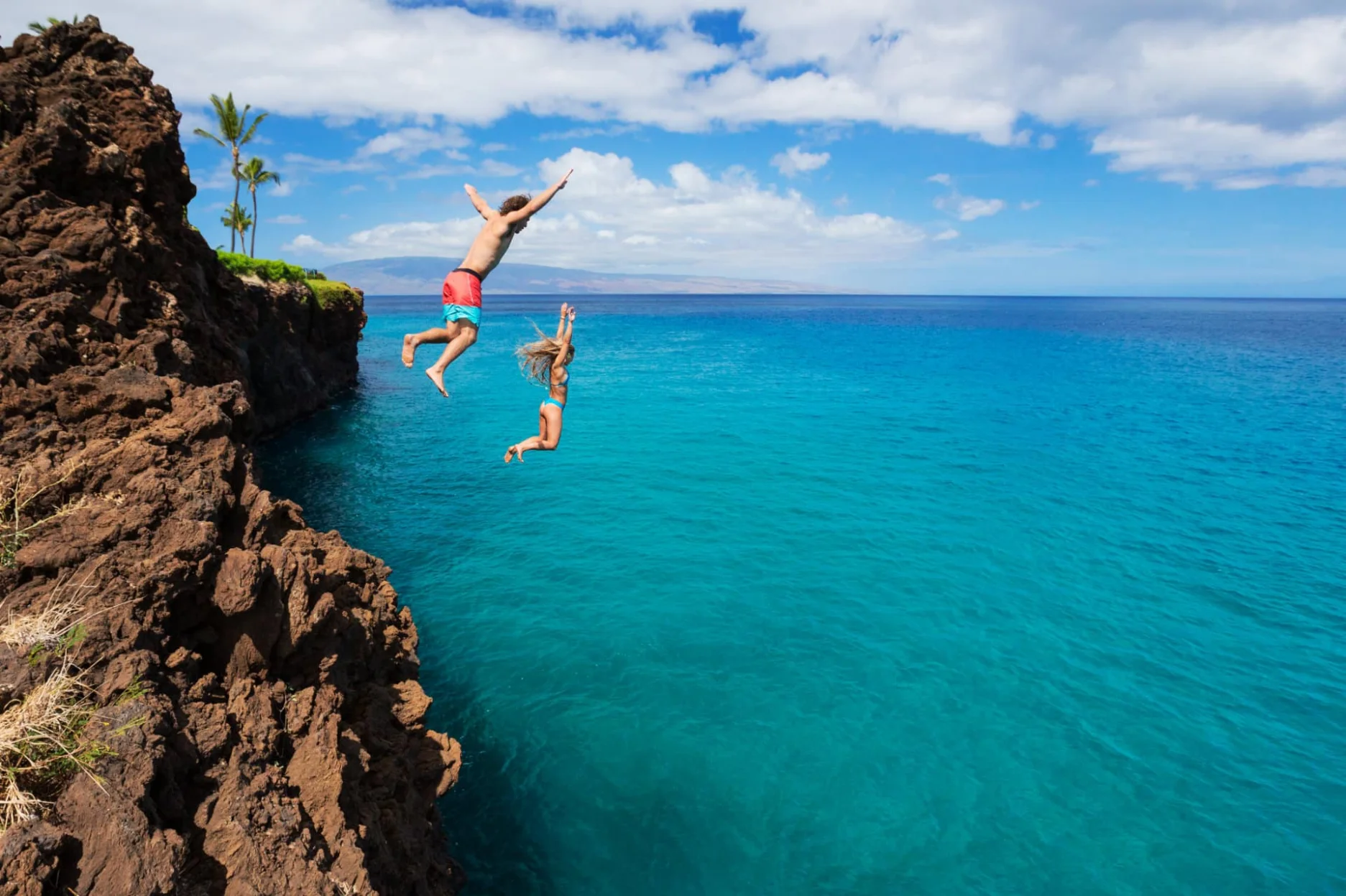 Two individuals jumping off a rocky cliff into vibrant turquoise waters below under a bright sky with fluffy clouds.